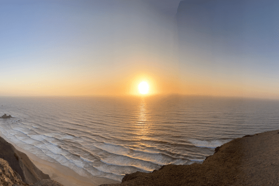 Panoramic view of sunset over ocean with cliffs and people on a rocky coastline.