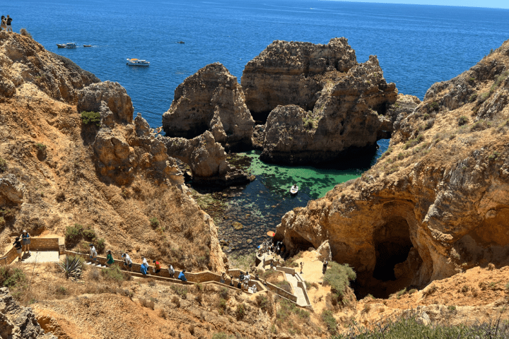 Rocky coastal cliffs with people on a winding path, clear blue sea and boats in the background.