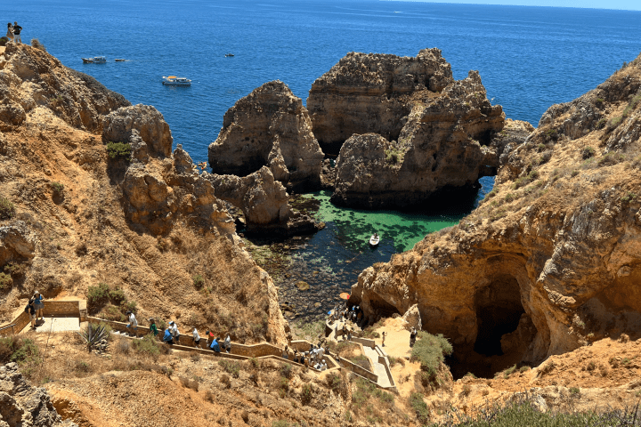 People walking on path through rocky cliffs by the ocean with small boats in the water.