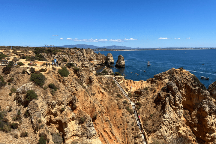 Rocky coastal landscape with cliffs, stairs, ocean, and people walking under a clear blue sky.