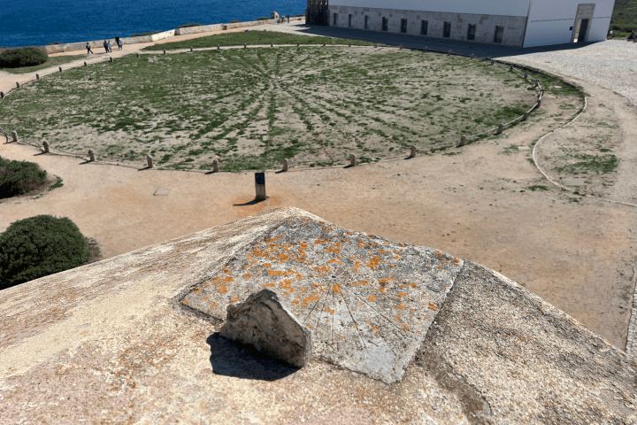 Sundial on stone ledge overlooking circular grassy area and blue sea.