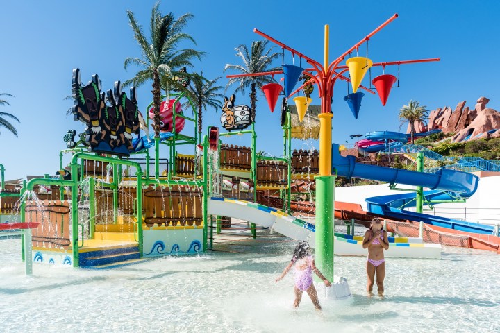 Children play in a colorful water park with slides and palm trees under a clear sky.