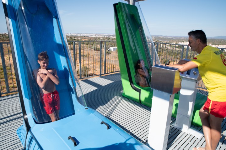 Two people in vertical water slide capsules, one in blue and one in green, with an operator assisting nearby.