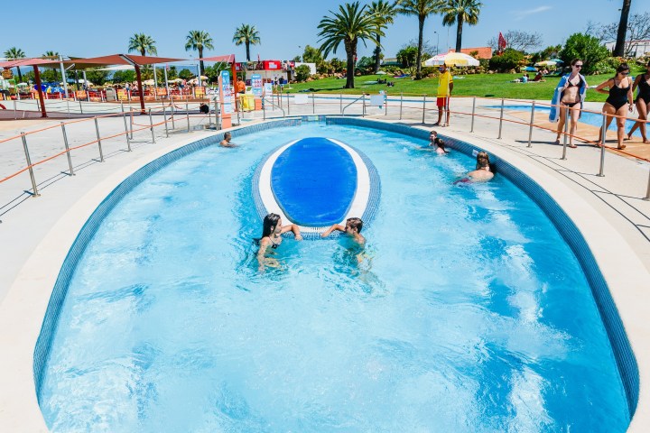 People enjoying an oval-shaped pool with palm trees and umbrellas in the background.