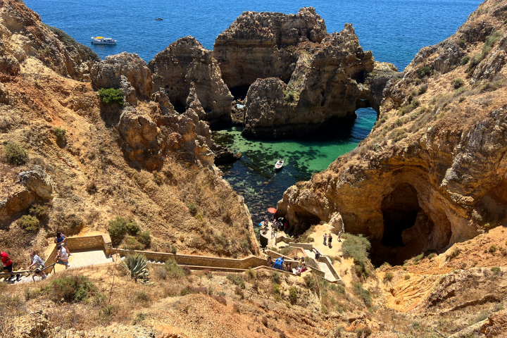 Rocky coastal landscape with cliffs, caves, clear water, boats, and people on stairs.
