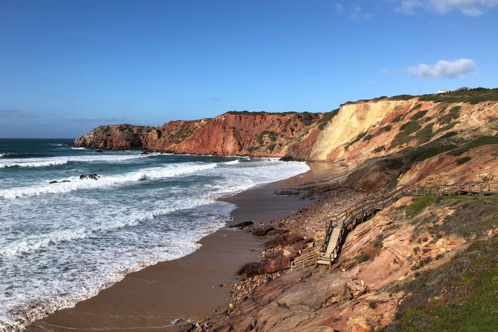 Coastal cliff with waves, sandy beach, and footpath under a clear blue sky.