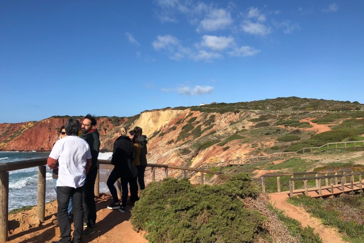 Group of people standing by a coastal cliff railing with blue sky and ocean waves.