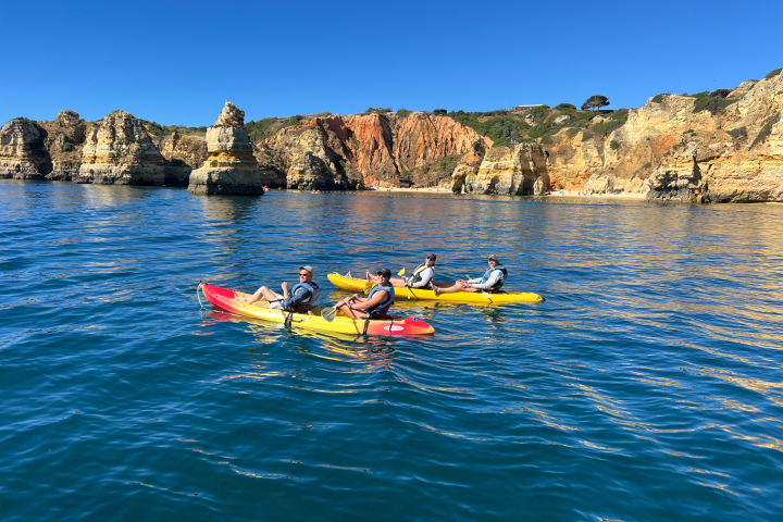 Four people kayaking on a blue sea near rocky cliffs under a clear blue sky.