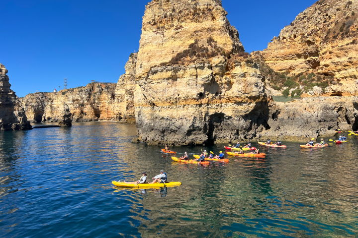 Group of kayakers paddling near towering cliffs under a clear blue sky.