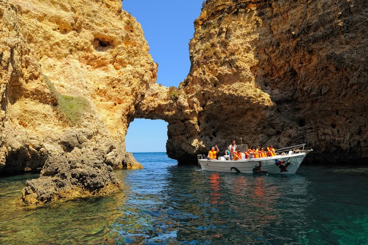 Boat with people in life jackets near rocky arch on a sunny day.