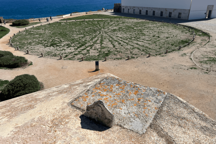 Circular grassy area with stone path and sea view, bordered by a building and blue sky.