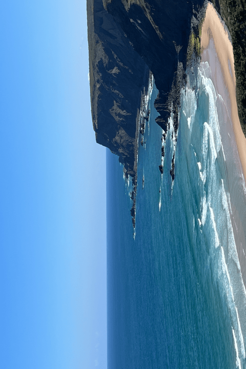 Scenic view of a coastal cliff with waves crashing on a sandy beach and clear blue sky.