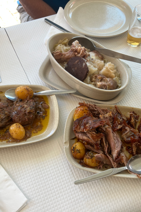 Table with dishes of meat, potatoes, and olives, with utensils and plates.