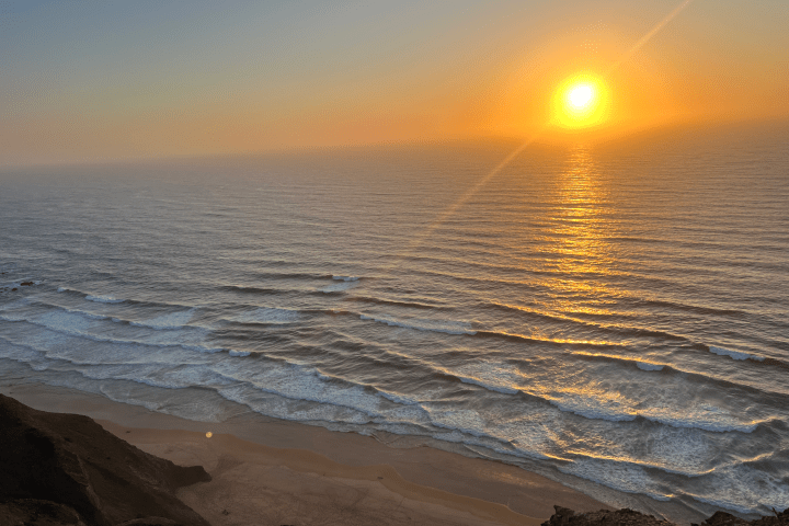 Sun setting over ocean with orange sky and gentle waves reaching a rocky beach.
