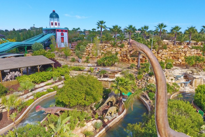 Amusement park with dinosaur statues, water ride, and greenery under a clear sky.