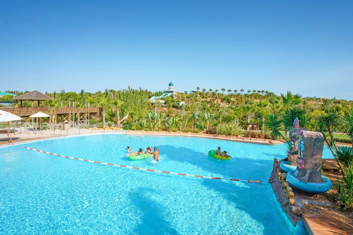 People in a large pool with inflatables, surrounded by tropical plants and water slides in the background.